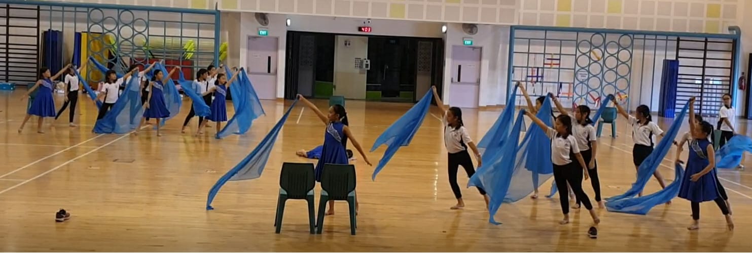 Girls perform a dance routine in a school hall, waving large blue fabric pieces while wearing blue and white outfits.